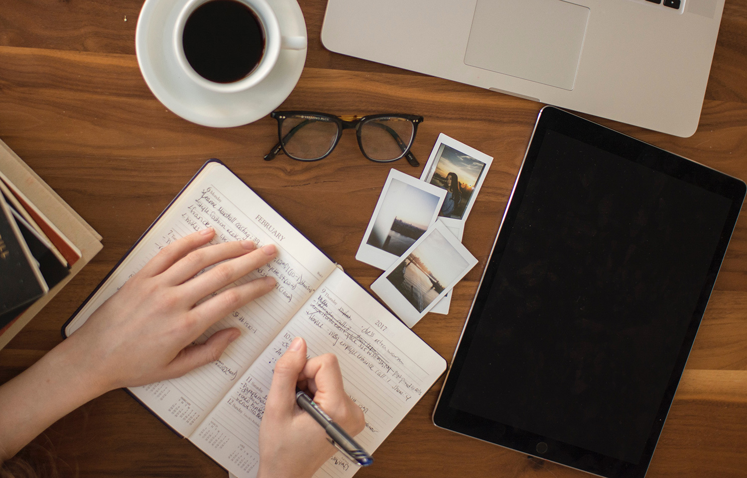 A person writes in a notebook on a desk strewn with photos, a notebook, a laptop, glasses, and a cup of coffee