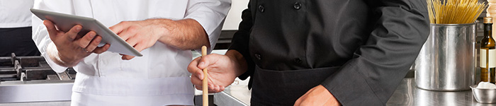 A chef in a white jacket shows an iPad to a chef in a black jacket, who is stirring a dish.