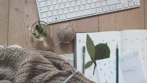 A keyboard, a notebook, a ball of yarn, and a crochet work in progress on a wooden desk.