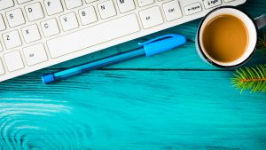 A keyboard sitting on a bright blue wooden surface with a ballpoint pen, a mug of milky coffee, and a pine branch