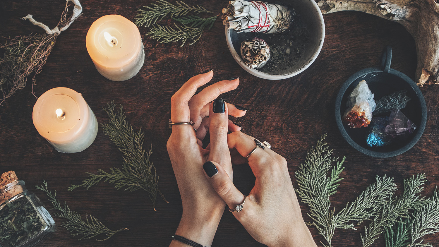 A person's hands are resting on a table, with candles, tree branches, crystals, and other witchy accoutrements.