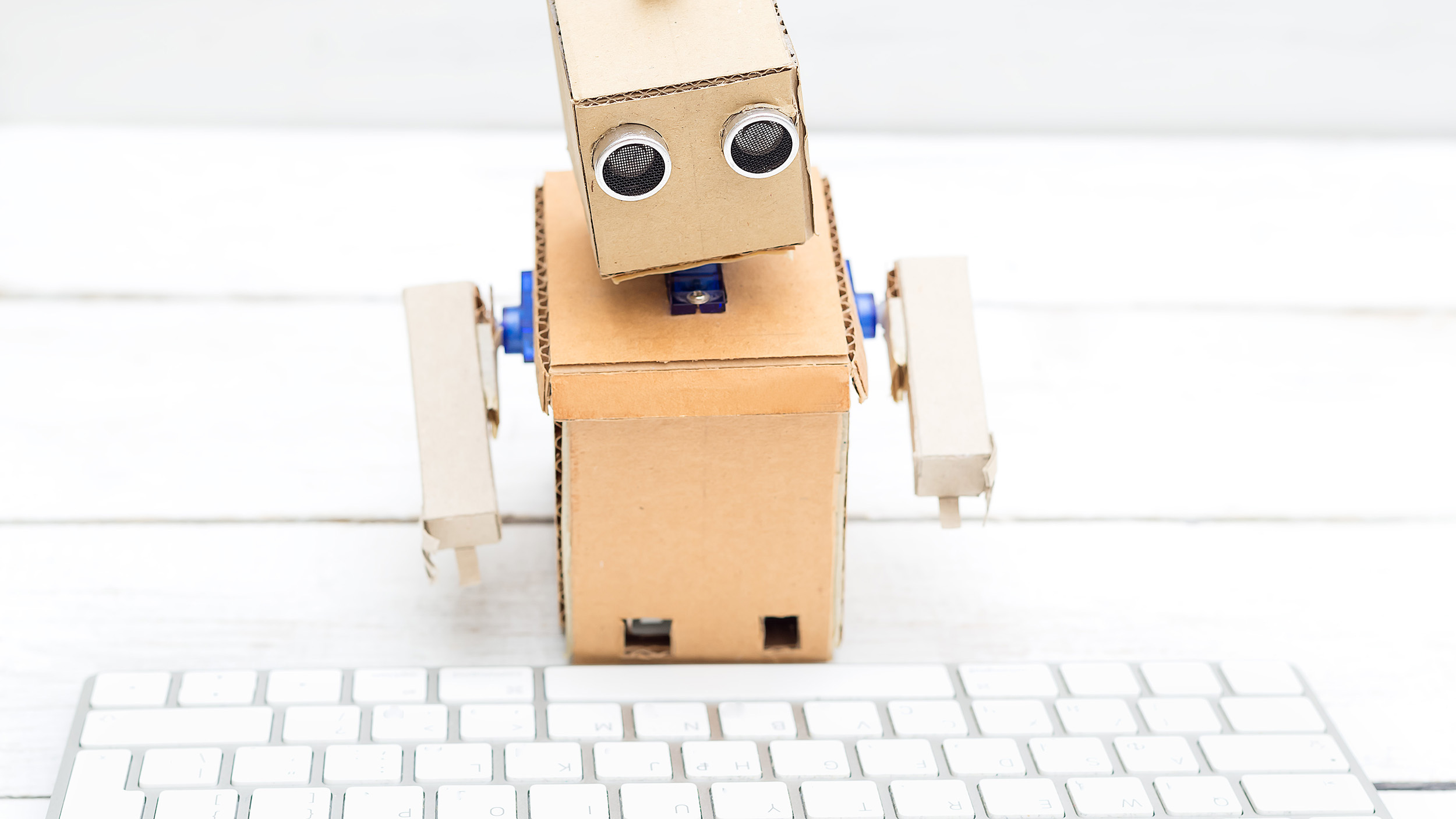 A small robot made of plywood sits on a white plank surface and is facing a white keyboard.